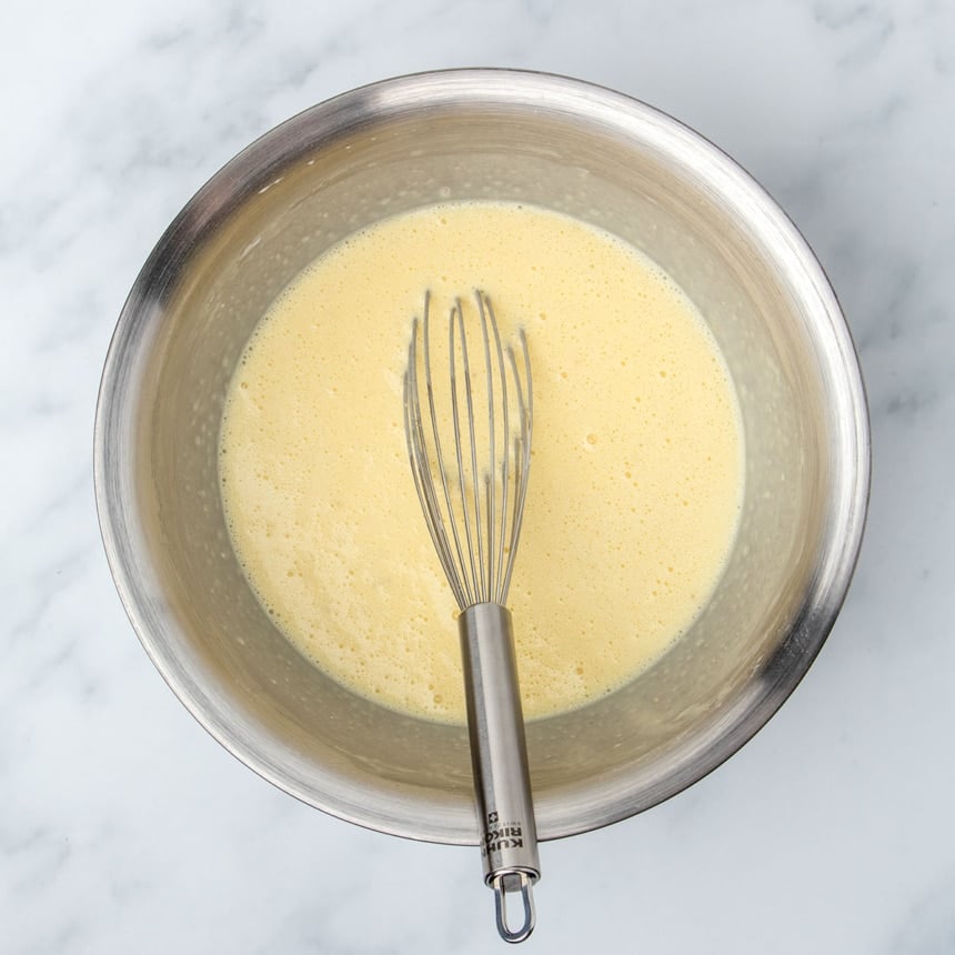 large round metal mixing bowl with frothy light yellow liquid mixture and metal wire whisk