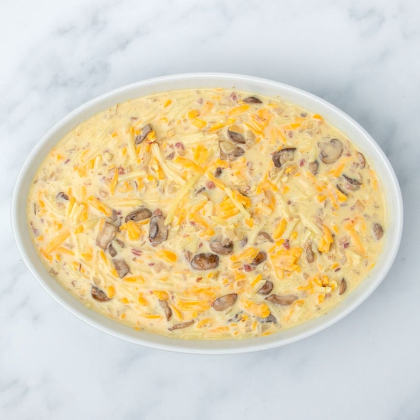 same raw breakfast casserole yellow liquid mixture with brown, yellow and pink pieces now in white oval casserole dish