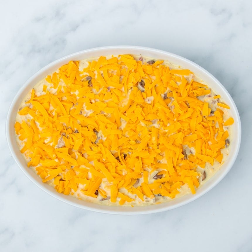 same casserole dish now with orange shredded cheddar cheese scattered on top of raw mixture