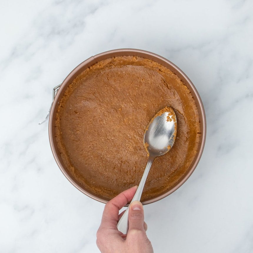 hand holding metal mixing spoon pressing brown graham cracker crust into even layer in gold spring form pan