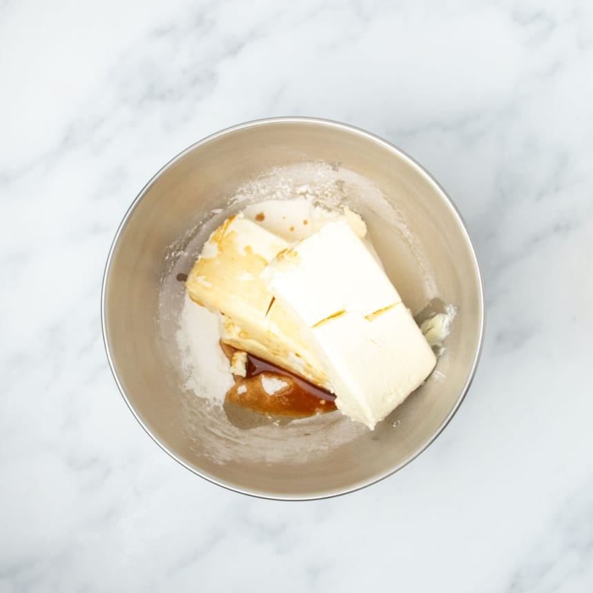 round metal stand mixer bowl with 2 blocks of white cream cheese, white starch, and brown liquid vanilla extract