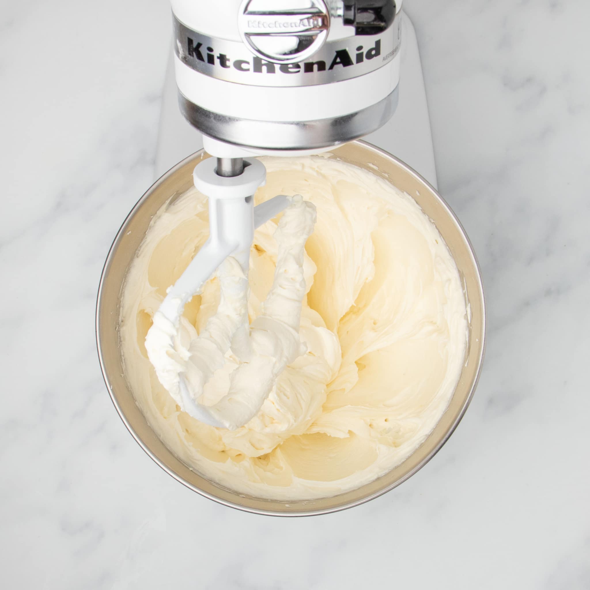 same mixing bowl with cream colored fluffy mixture of beaten cream cheese with white paddle attachment and white kitchenaid mixer