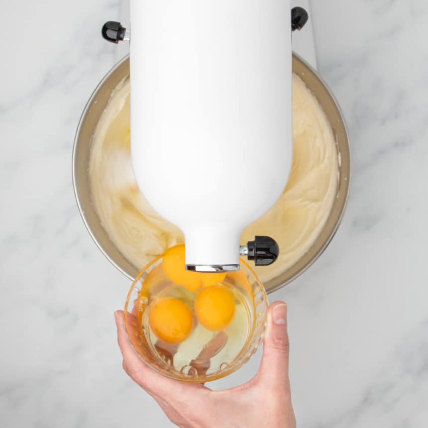 hand holding small glass bowl pouring 3 raw eggs into white kitchen aid mixer with mixer turned on