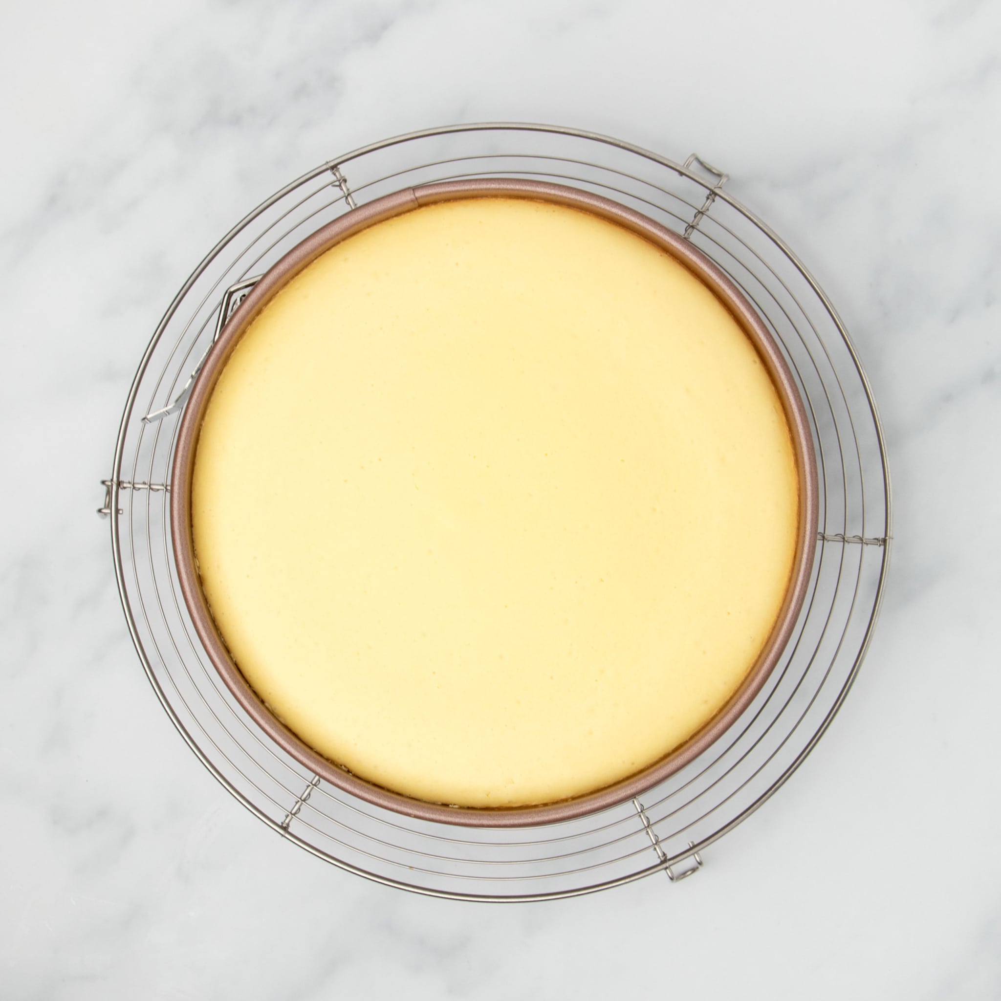 same cheesecake mixture in gold springform pan now baked into slightly darker yellow cake on round wire rack