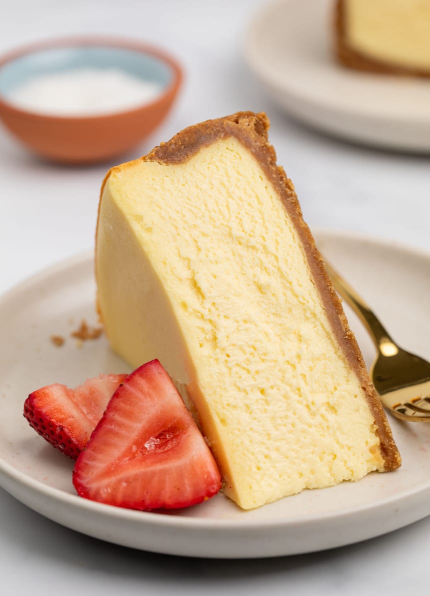 triangular slice of yellow cheesecake with brown crust sitting on its side on a small cream colored plate with two red strawberry halves and a gold colored small fork