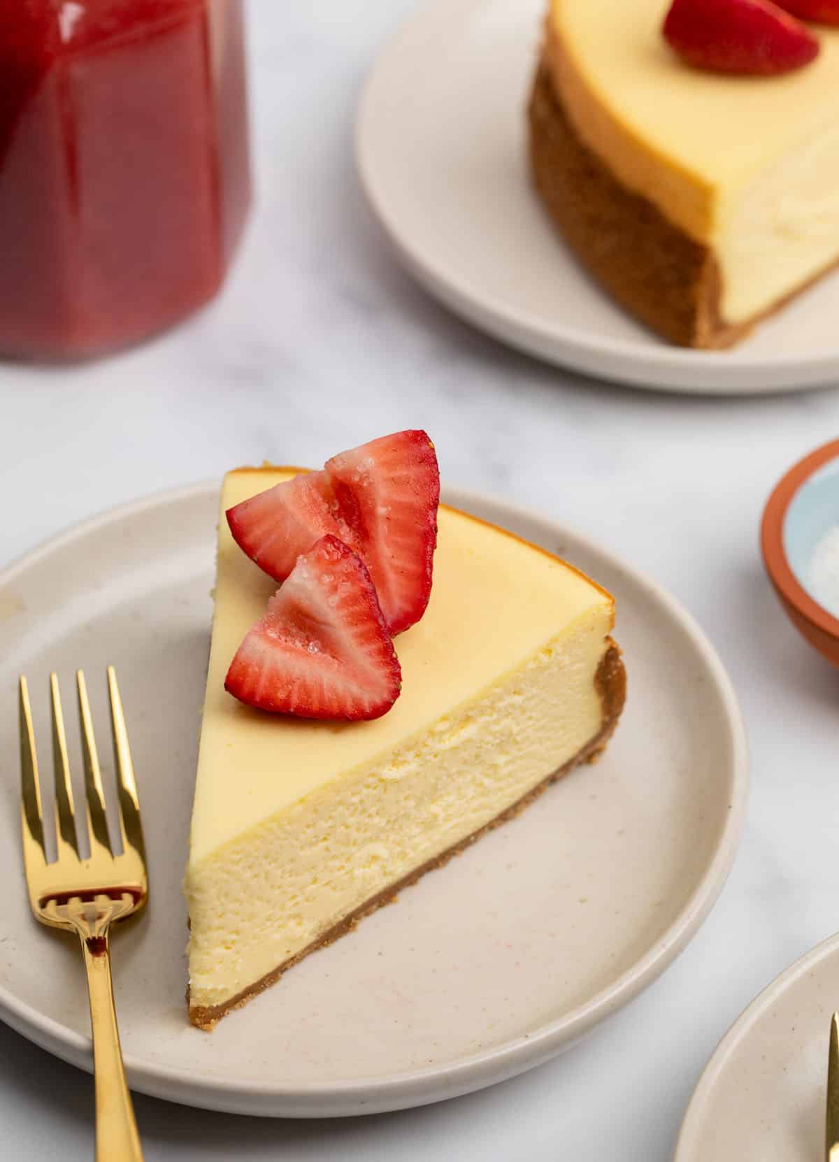 triangular yellow baked cheesecake slice with two halves of red strawberry with some grains of coarse sugar on top on small cream plate with gold colored small fork and more plated cheesecake in background