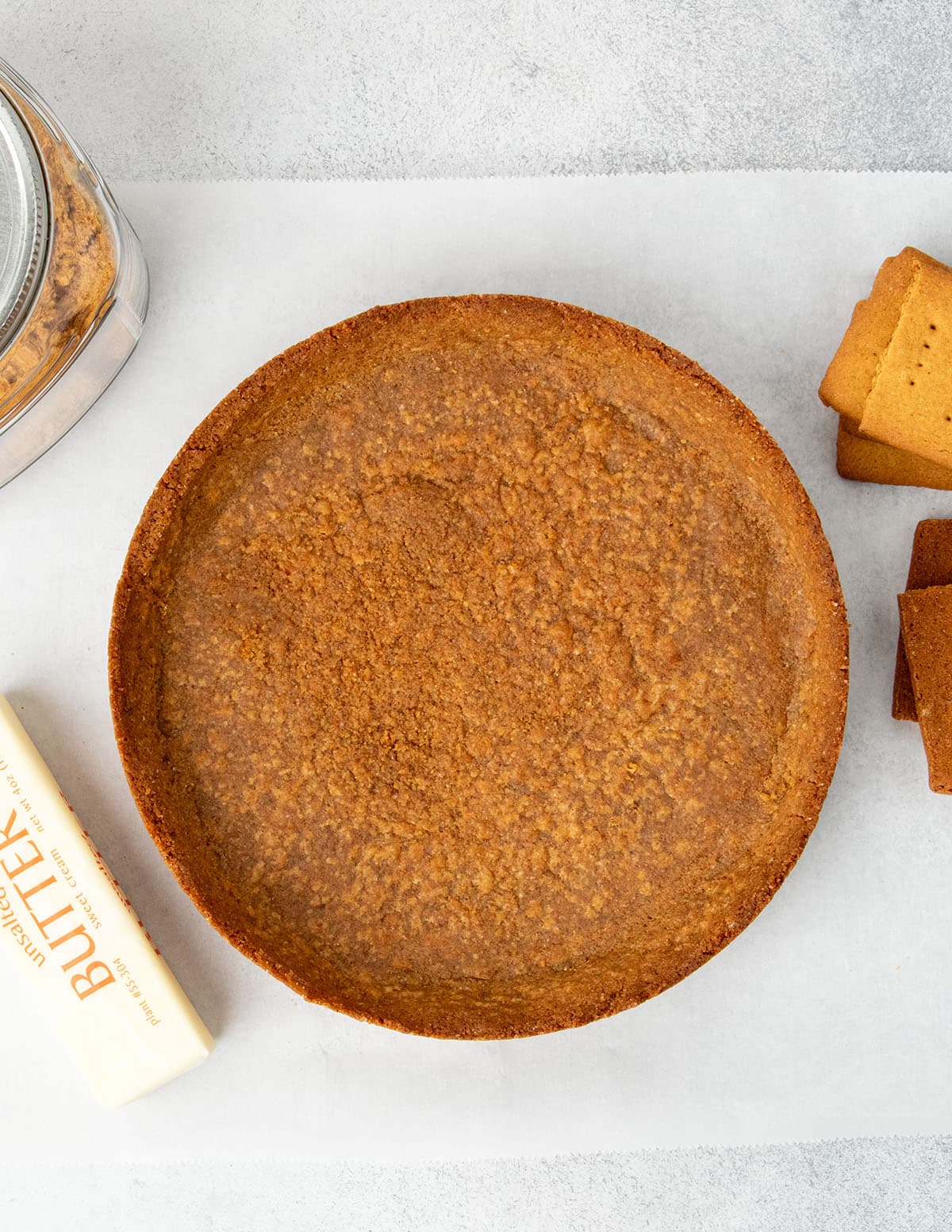 Brown baked graham cracker crust on white paper with a stick of butter, more graham crackers and a glass jar with cracker crumbs.