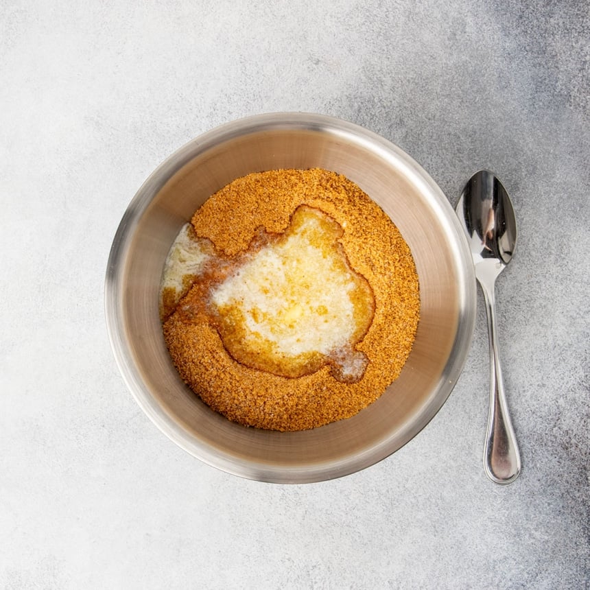 same bowl with brown crumbs whisked with white sugar and melted yellow butter sitting on top with a spoon next to the bowl