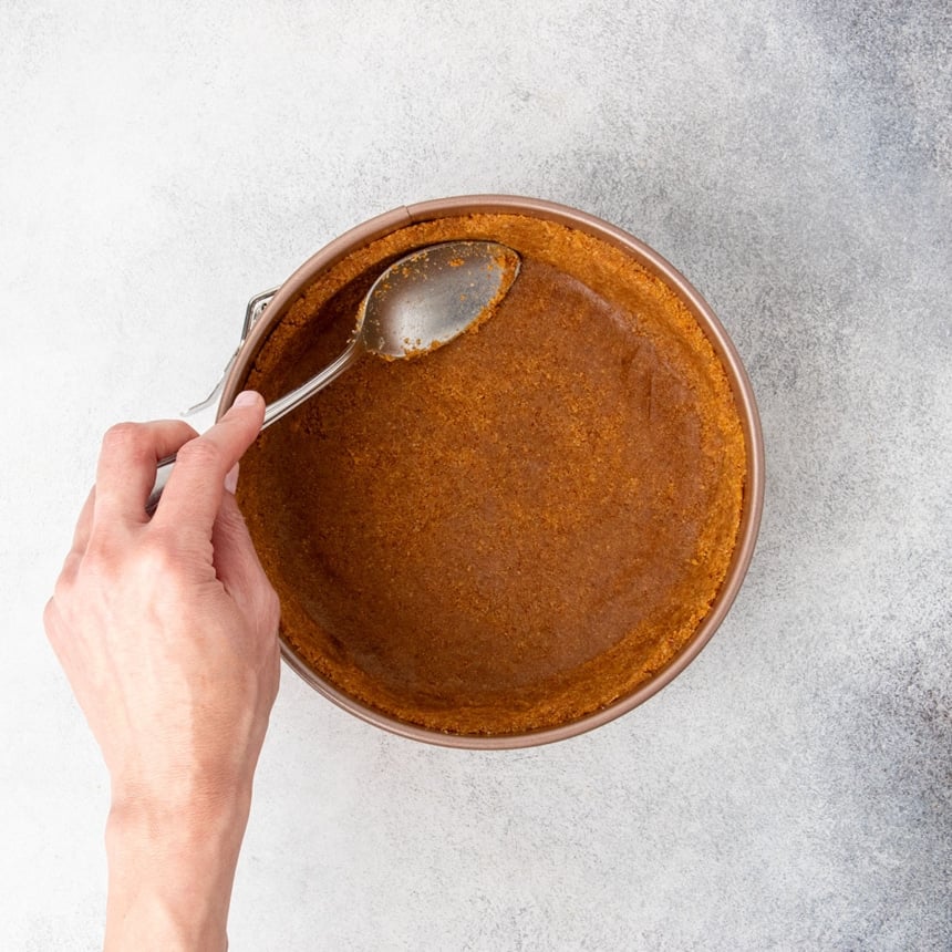 hand using large metal mixing spoon to pat down brown clumpy crumb mixture into bottom and up sides of pan