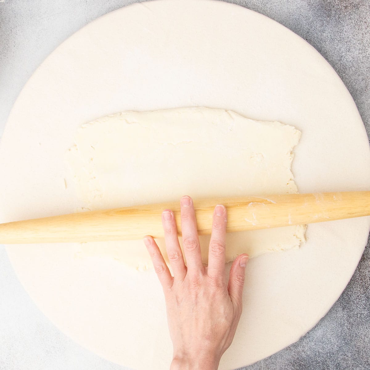 Fingers rolling same blonde rolling pin on top of rectangle or raw dough on board.