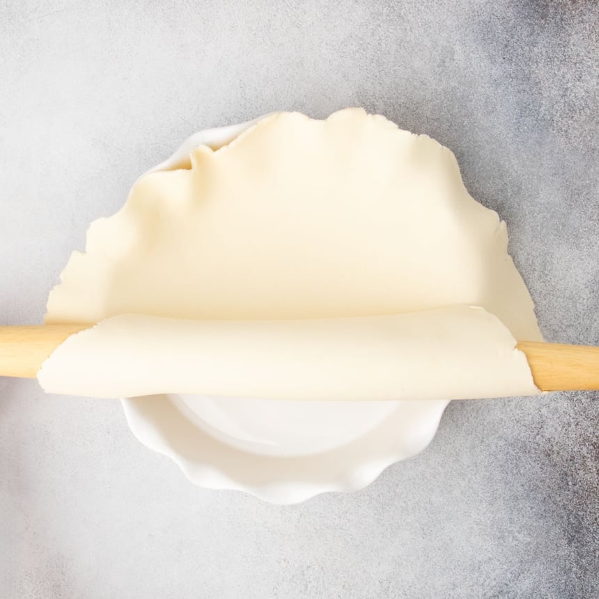 rolled round of raw pie crust being rolled off of blonde wood rolling pin onto white porcelain scalloped edge pie dish