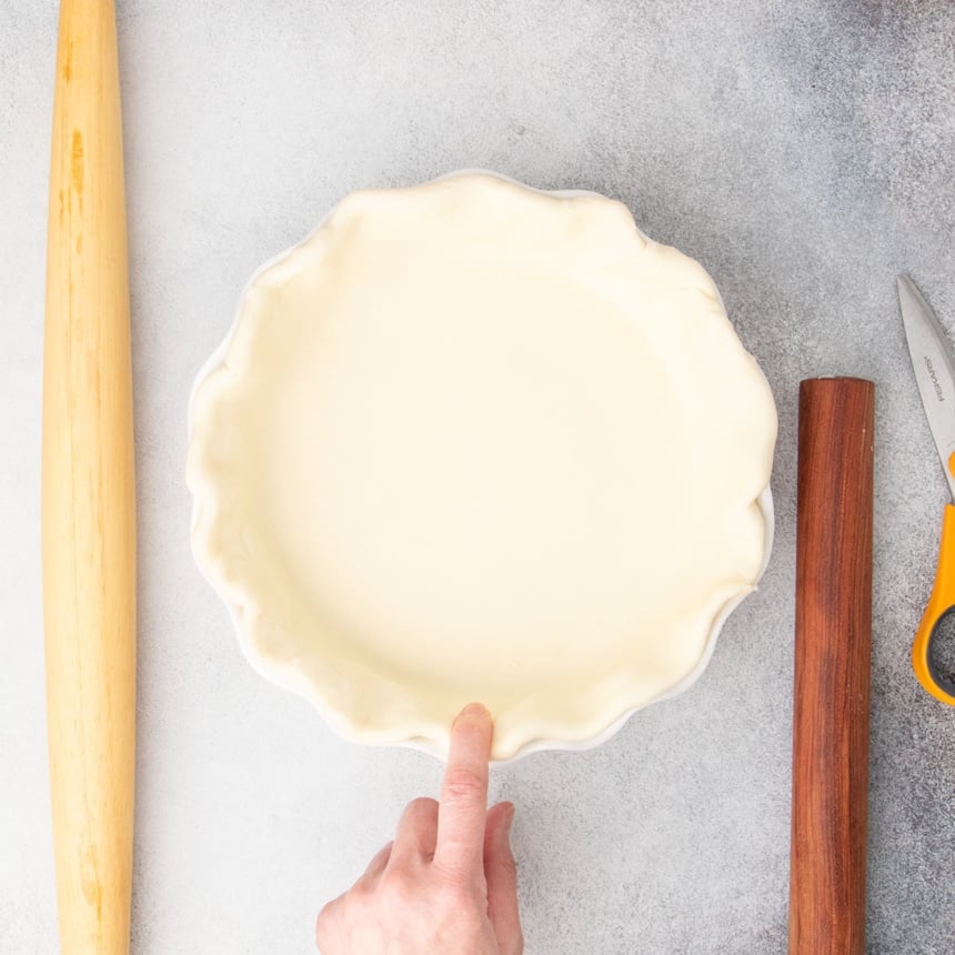 finger pressing edge of shaped pie raw pie crust in white pie dish with two rolling pins and kitchen shears nearby on light gray surface