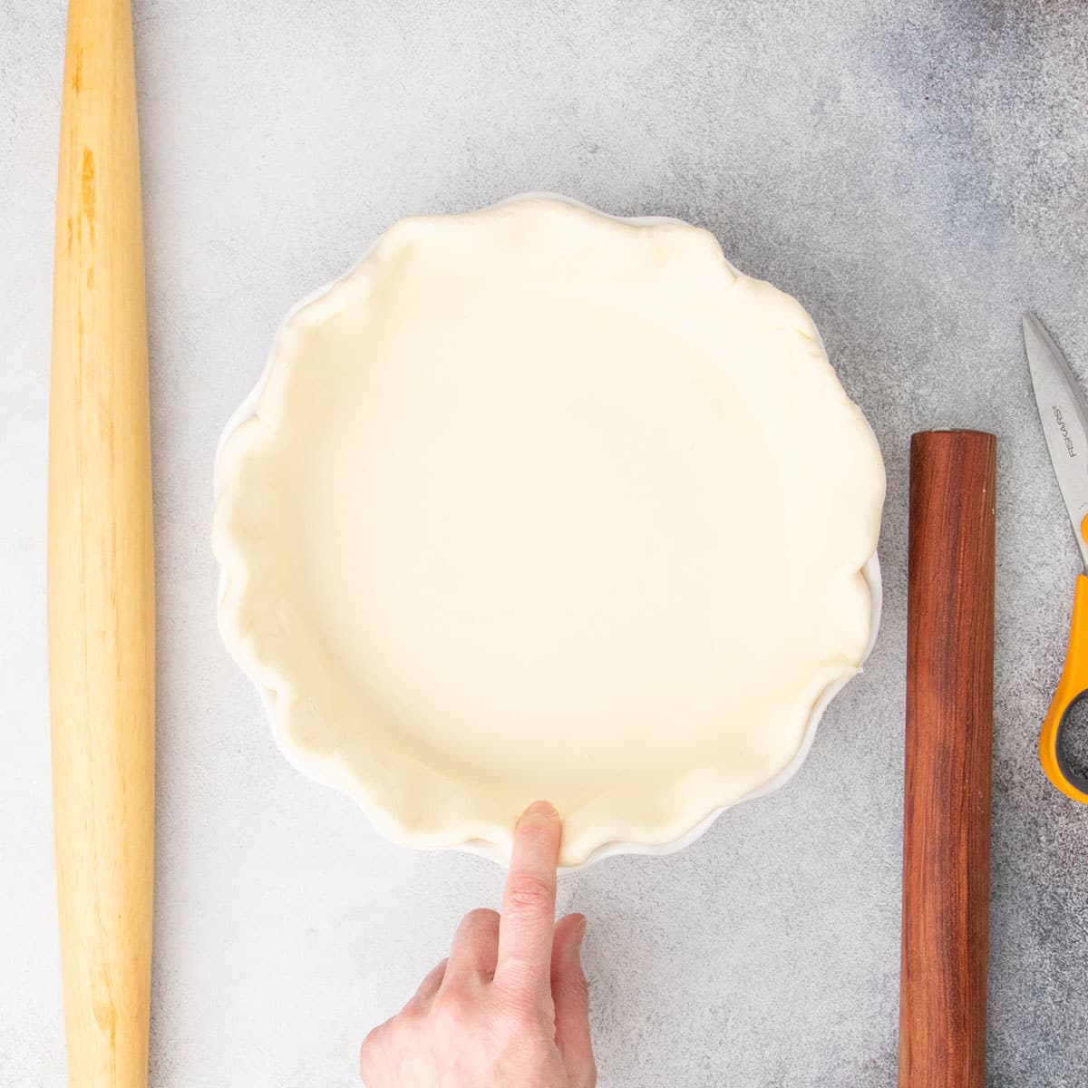 finger pressing edge of shaped pie raw pie crust in white pie dish with two rolling pins and kitchen shears nearby on light gray surface