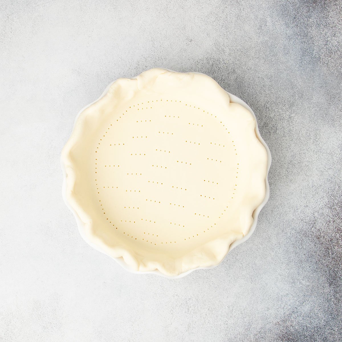 raw shaped pie crust in white pie dish with piercings from tines of fork all over bottom of crust