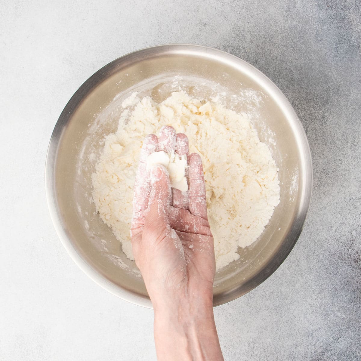 hand with flour on fingers pressing pieces of butter covered in flour flat between thumb and fingers, held over mixing bowl