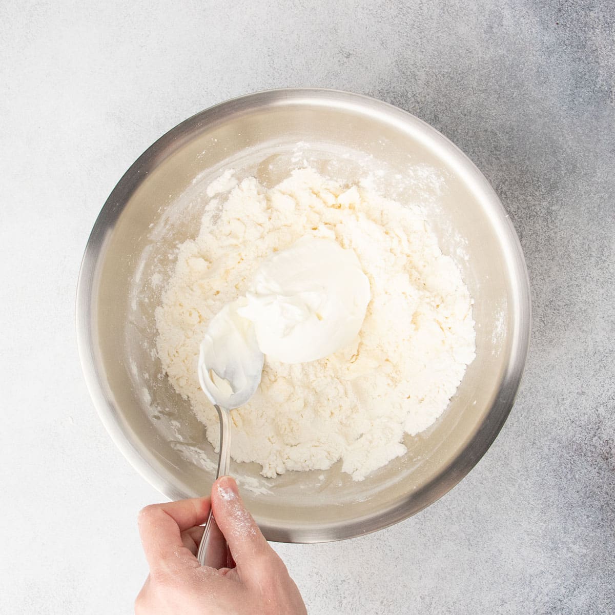 Hand holding large metal mixing spoon placing sour cream in mixing bowl on top of flour and butter mixture.