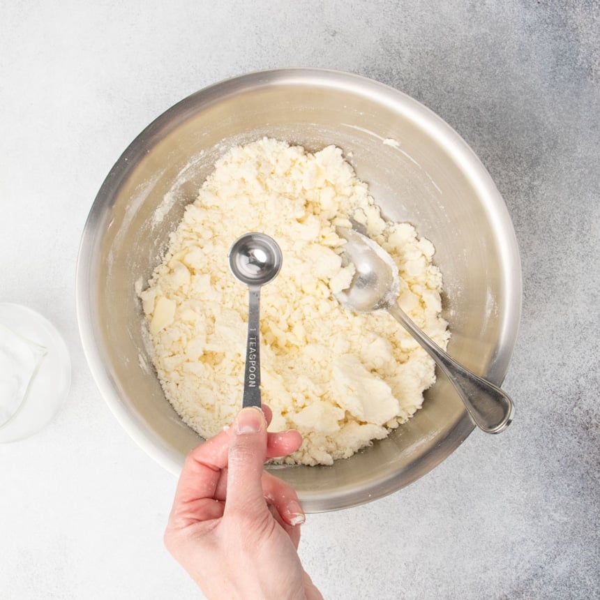 hand holding metal teaspoon with water about to be poured into mixing bowl with slightly dry somewhat clumpy light tan pastry mixture