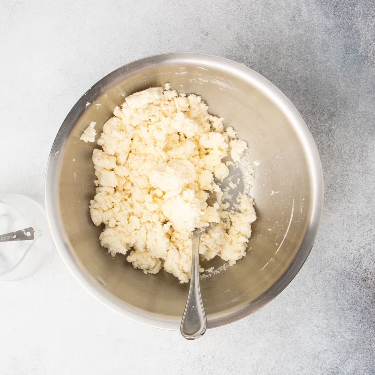 Light tan clumpy pie crust mixture in same mixing bowl with metal mixing spoon.