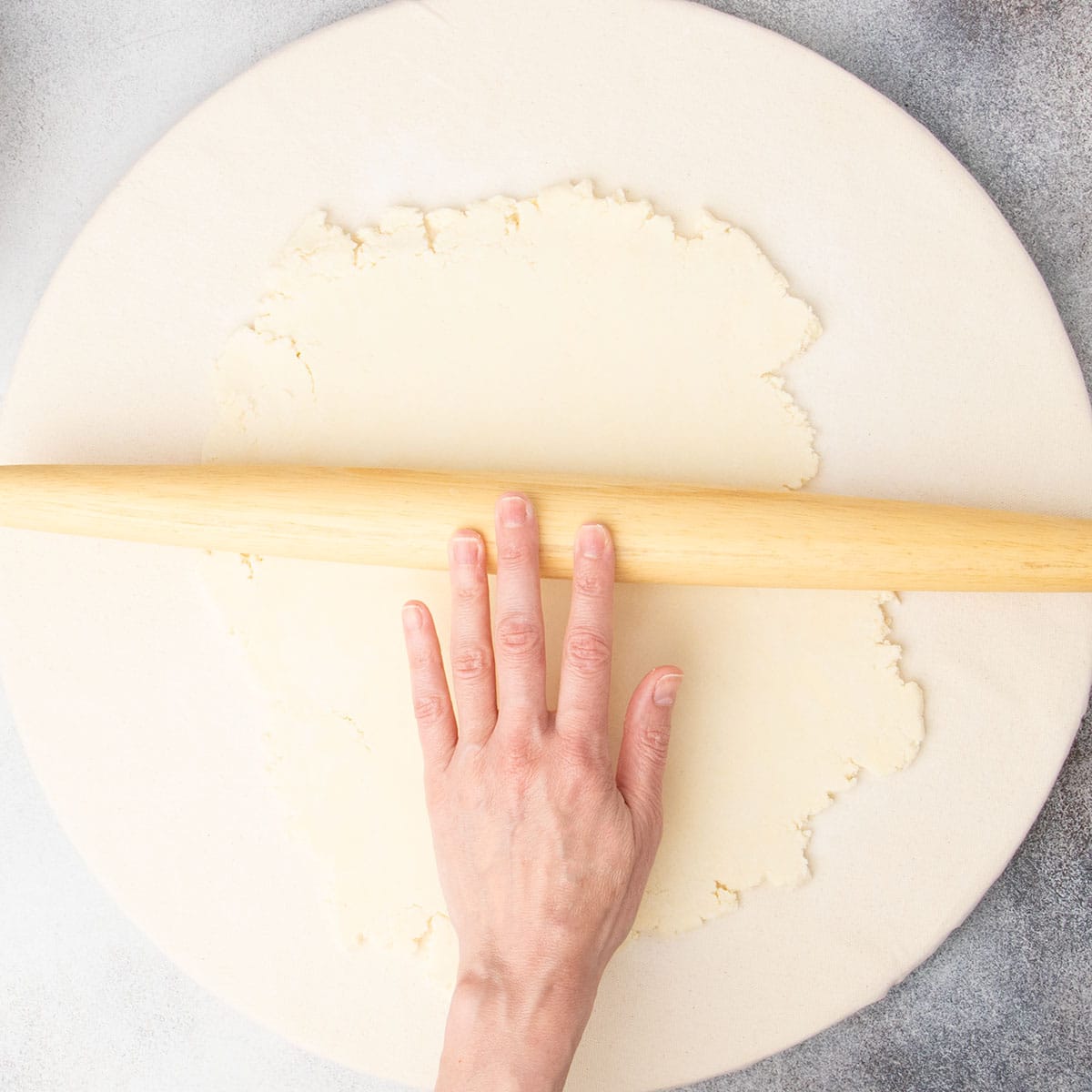 fingers rolling blonde wood tapered rolling pin to roll out light tan pie crust dough into rough rectangle on canvas covered pastry board