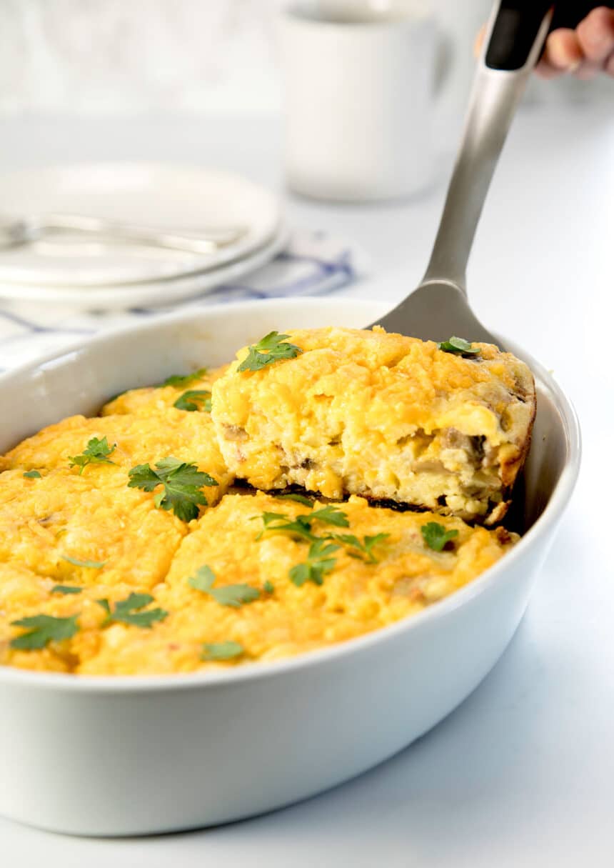 metal spatula picking up rectangular yellow slice of breakfast casserole with brown mushrooms and green parsley leaves from white oval casserole dish
