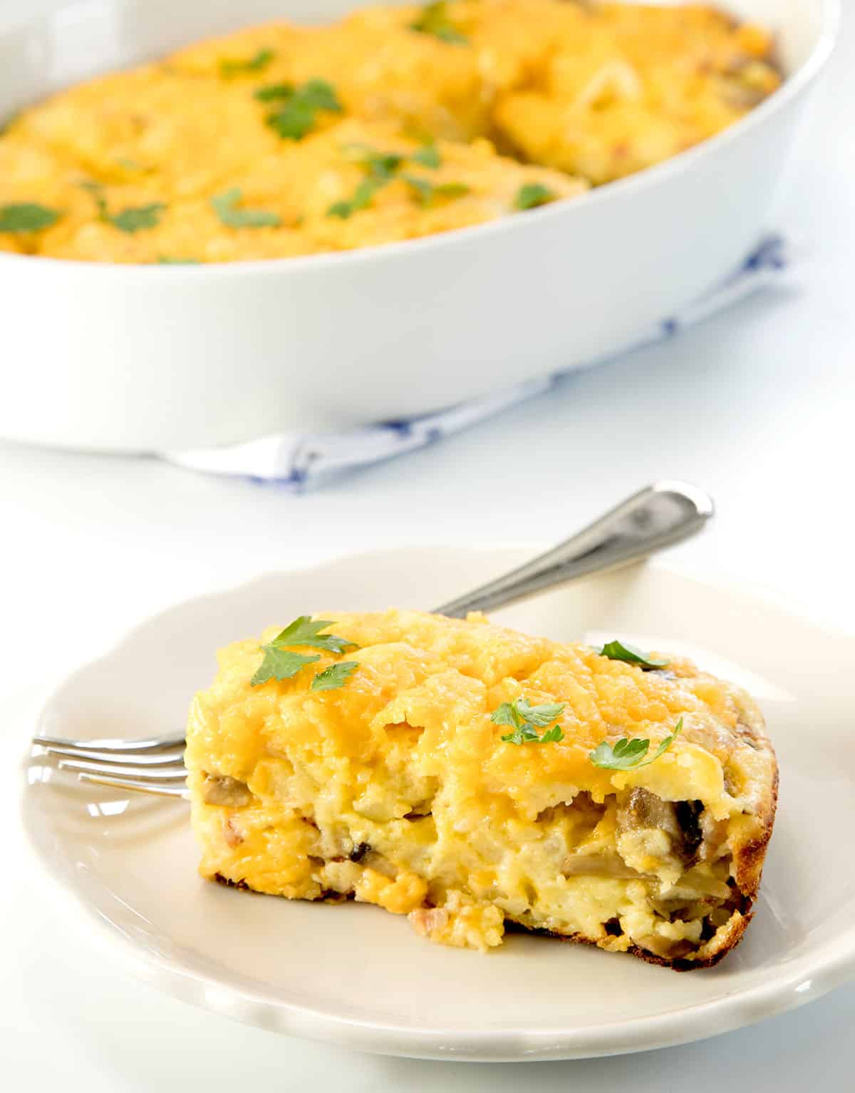 yellow rectangular slice of breakfast casserole with mushrooms inside and green parsley leaves on top on small white plate with small metal fork