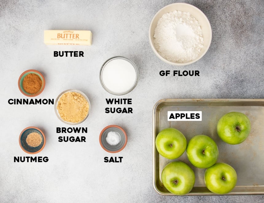 Overhead view of apples, gluten free flour, sugars, butter, and spices arranged on a gray surface.