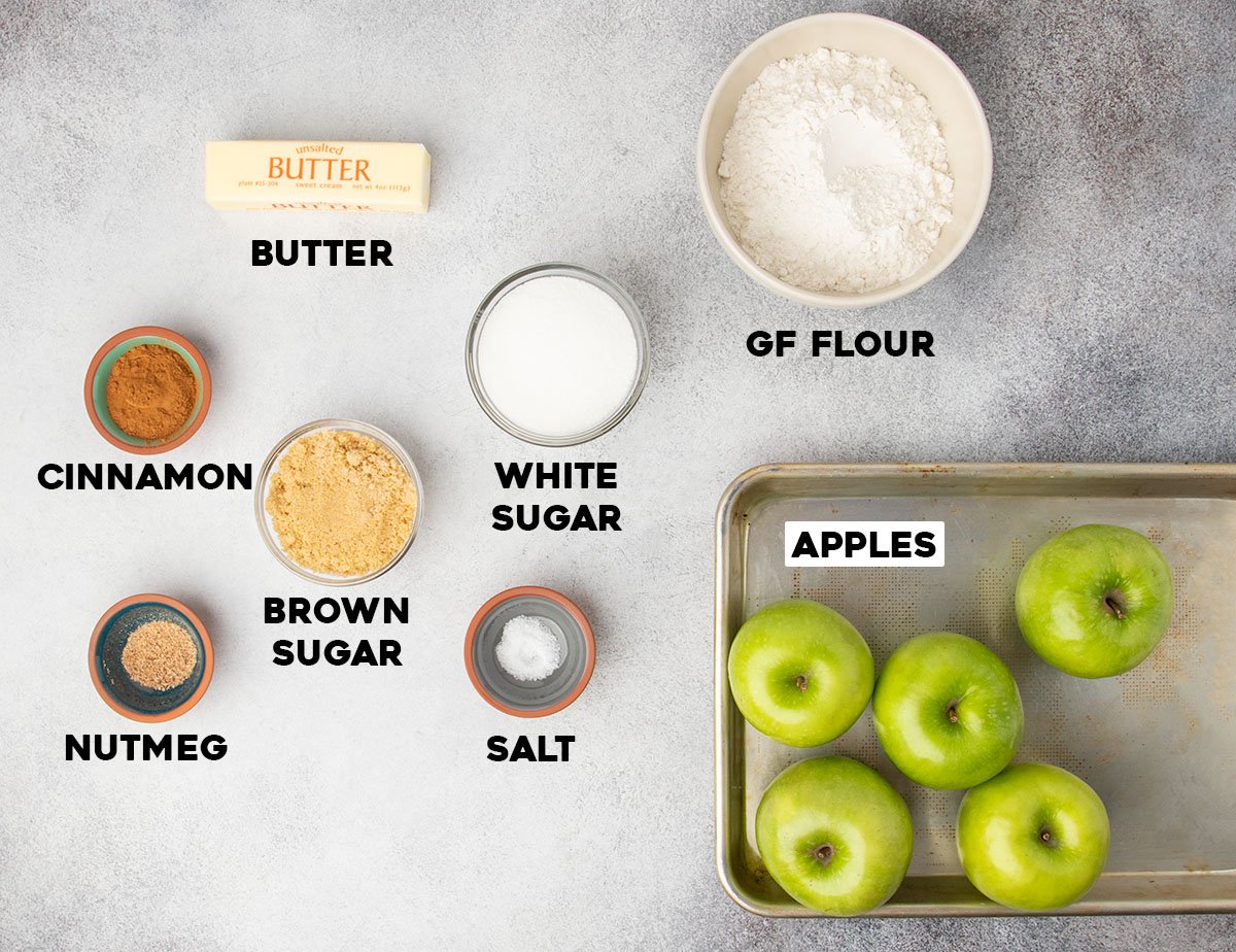 Overhead view of apples, gluten free flour, sugars, butter, and spices arranged on a gray surface.