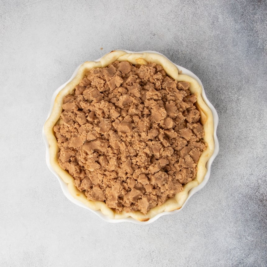 Unbaked apple pie filled with seasoned apples and topped with a crumb mixture with the edges of the parbaked pie crust visible along the perimeter.