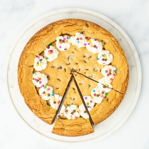 Top-down view of a decorated gluten free cookie cake with two slices cut, topped with piped frosting and colorful confetti sprinkles.