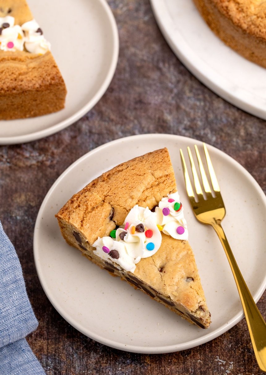 Single slice of cookie cake on a plate with piped frosting and rainbow sprinkles, next to a gold fork.