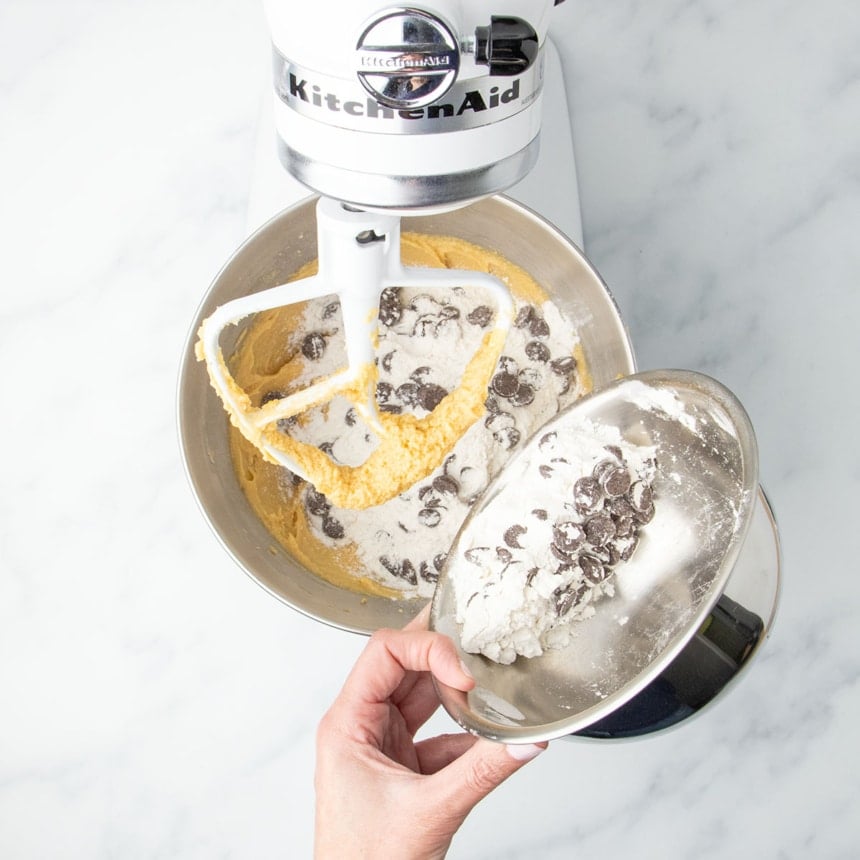 Hand tipping metal bowl with gluten free flour and chocolate chips into soft light brown dough in the mixing bowl with a stand mixer paddle attachment.
