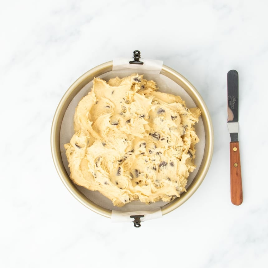 Unbaked raw chocolate chip cookie dough spread roughly in a gold colored round baking pan lined with parchment