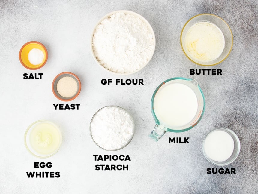 Overhead view of labeled ingredients for gluten free dinner rolls, including gluten free flour, tapioca starch, yeast, salt, sugar, egg whites, milk, and melted butter.