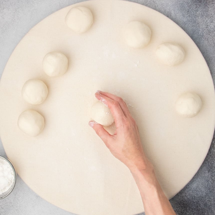 Hand shaping pieces of gluten free dough into round rolls on a floured surface.