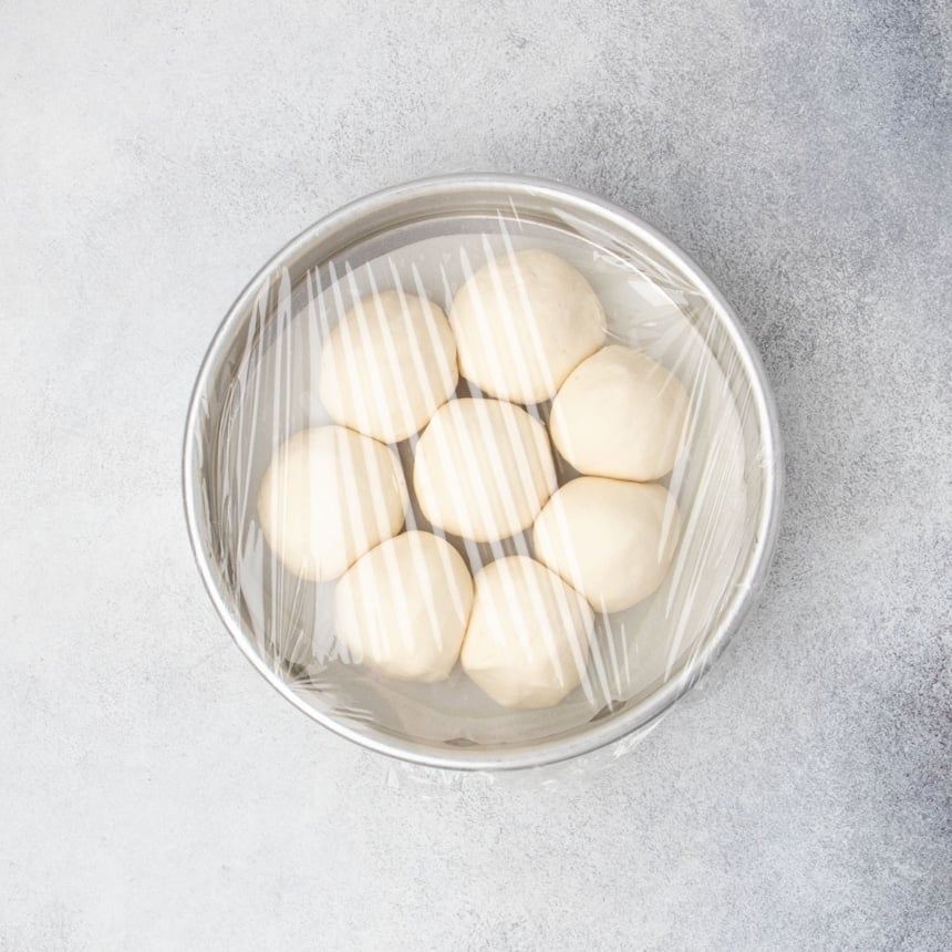Shaped raw round rolls arranged in a round baking pan, covered with plastic wrap.