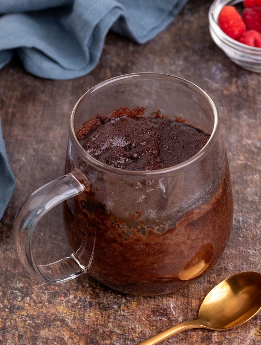 Freshly baked single-serve cake in a clear mug, with a soft, domed top and a gold spoon alongside.