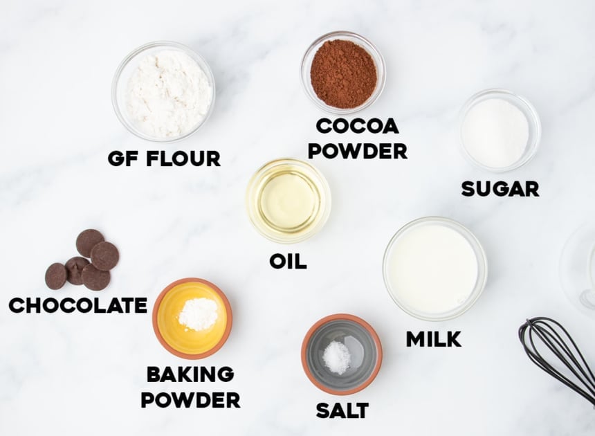 Ingredients for a gluten free mug cake, labeled and arranged on a white surface, including flour, cocoa, chocolate, oil, milk, and more