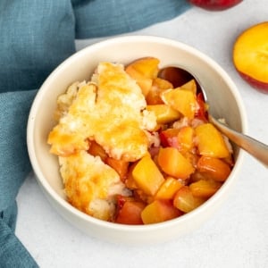 A cream colored bowl of warm chopped cooked peaches with fluffy golden topping, served with a spoon with a blue cloth and fresh peaches