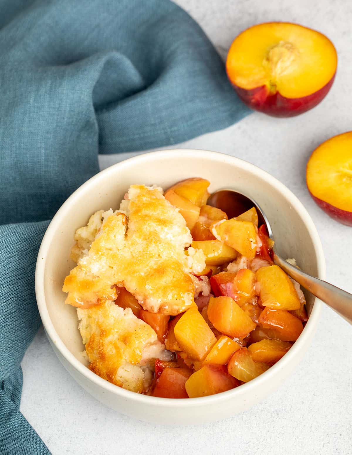 A bowl of warm gluten free peach cobbler with fluffy golden topping and juicy peach filling, served with a spoon.