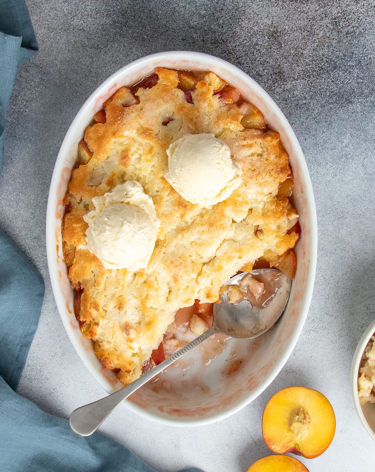 Peach cobbler in oval white serving dish served warm with scoops of vanilla ice cream melting into the golden topping
