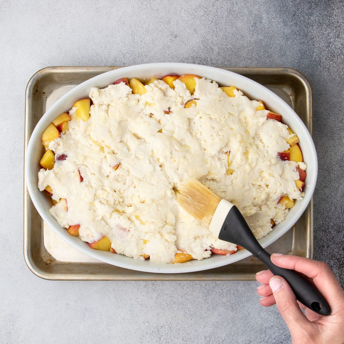 Raw, spread out cobbler topping being brushed with melted butter for a golden finish before baking.
