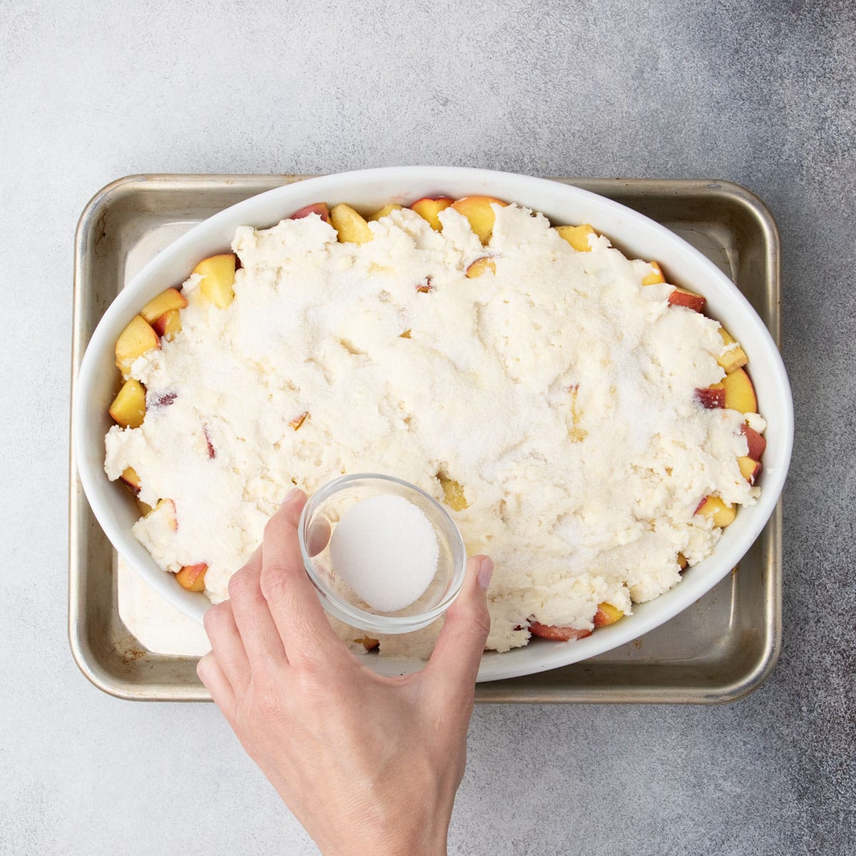 A hand sprinkles sugar over the cobbler dough to create a lightly crisp top.