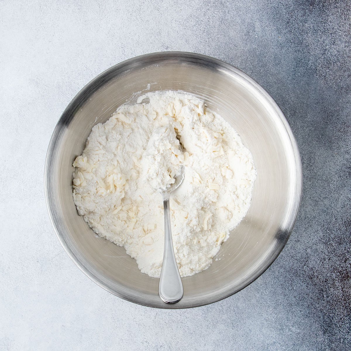 Gluten free flour and grated butter mixed together with a spoon in a metal bowl to form a loose mixture of cold grated butter in flour.