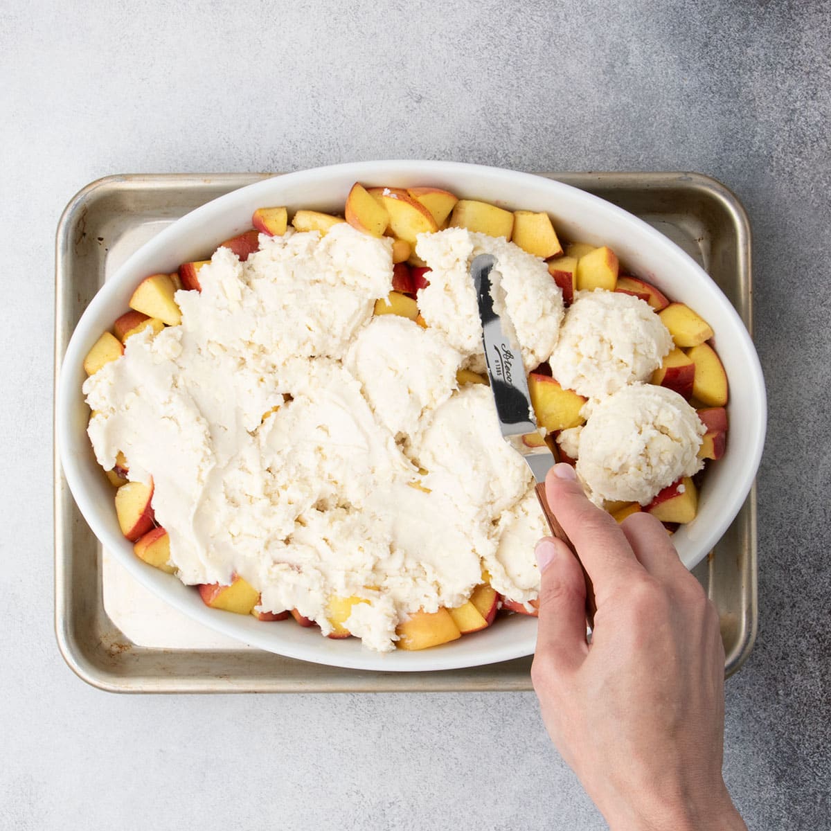 Hand using an offset spatula to spread cobbler dough evenly over the peach filling.