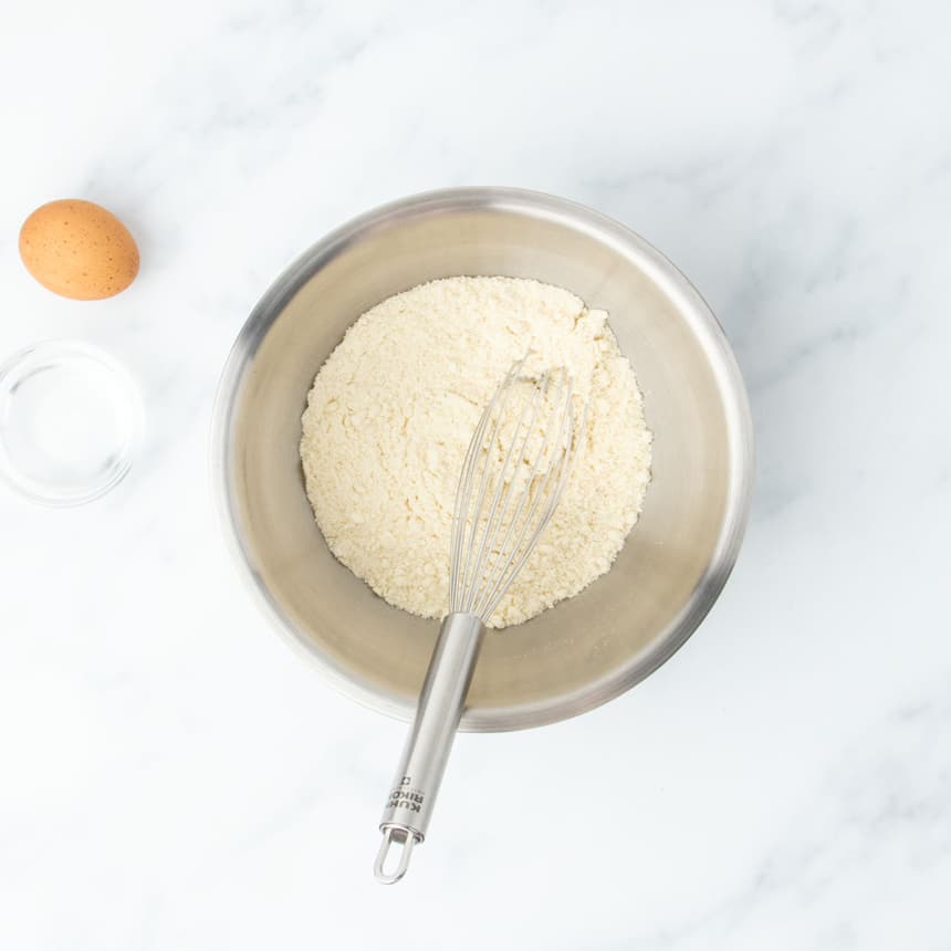 Bowl of almond flour with a whisk resting inside, egg and water nearby
