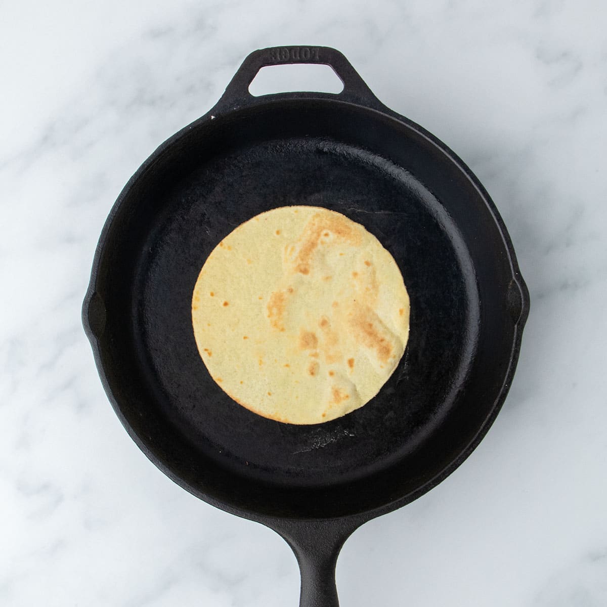 A single tortilla cooking in a black cast iron skillet on a marble surface.