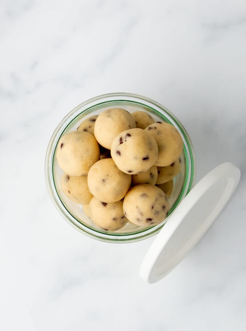 Top-down view of a glass jar filled with chocolate chip cookie dough balls, with the white plastic lid resting nearby on a light marble countertop.