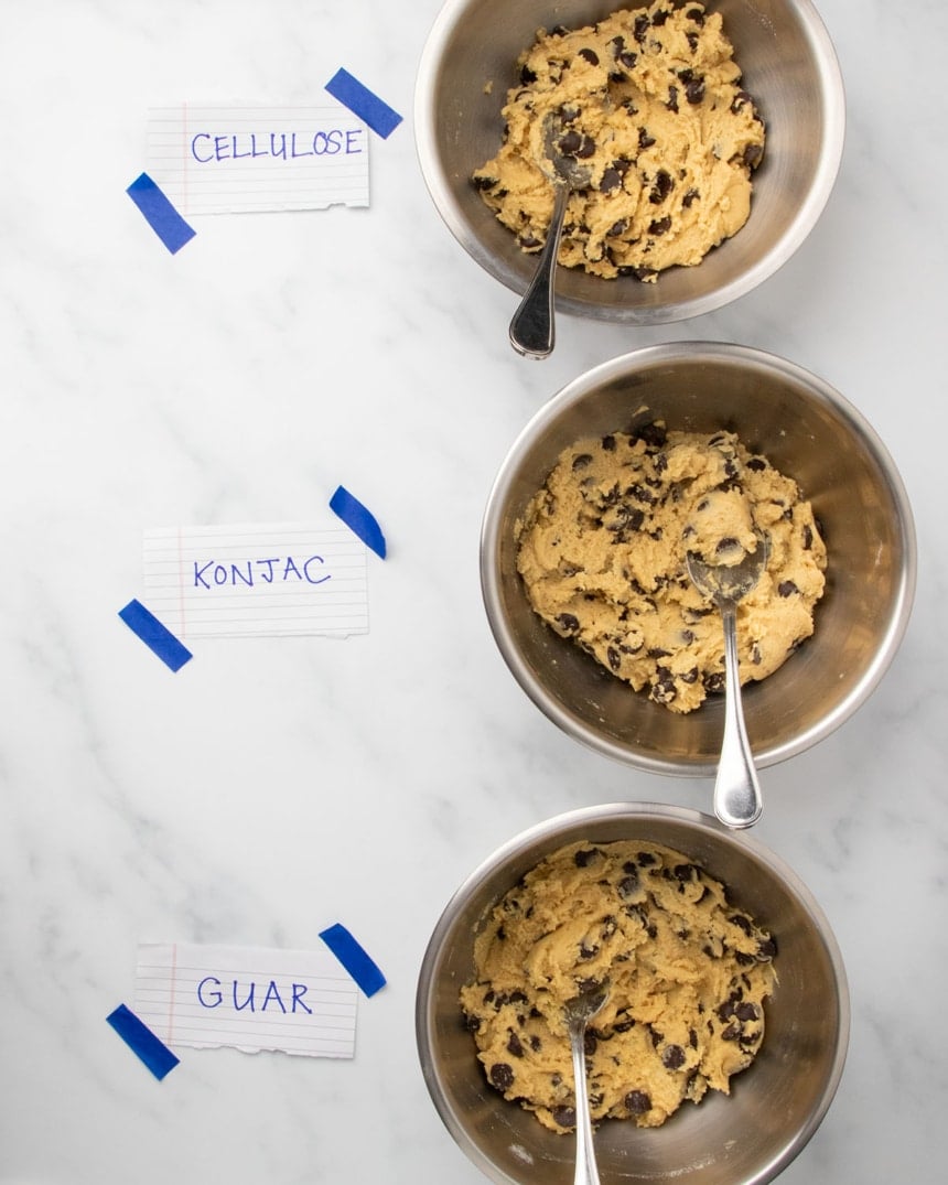 3 medium-size metal mixing bowls, each with raw unshaped gluten free chocolate chip cookie dough with a metal spoon, and each with a white paper with the word "cellulose" "konjac" or "guar" and blue tape