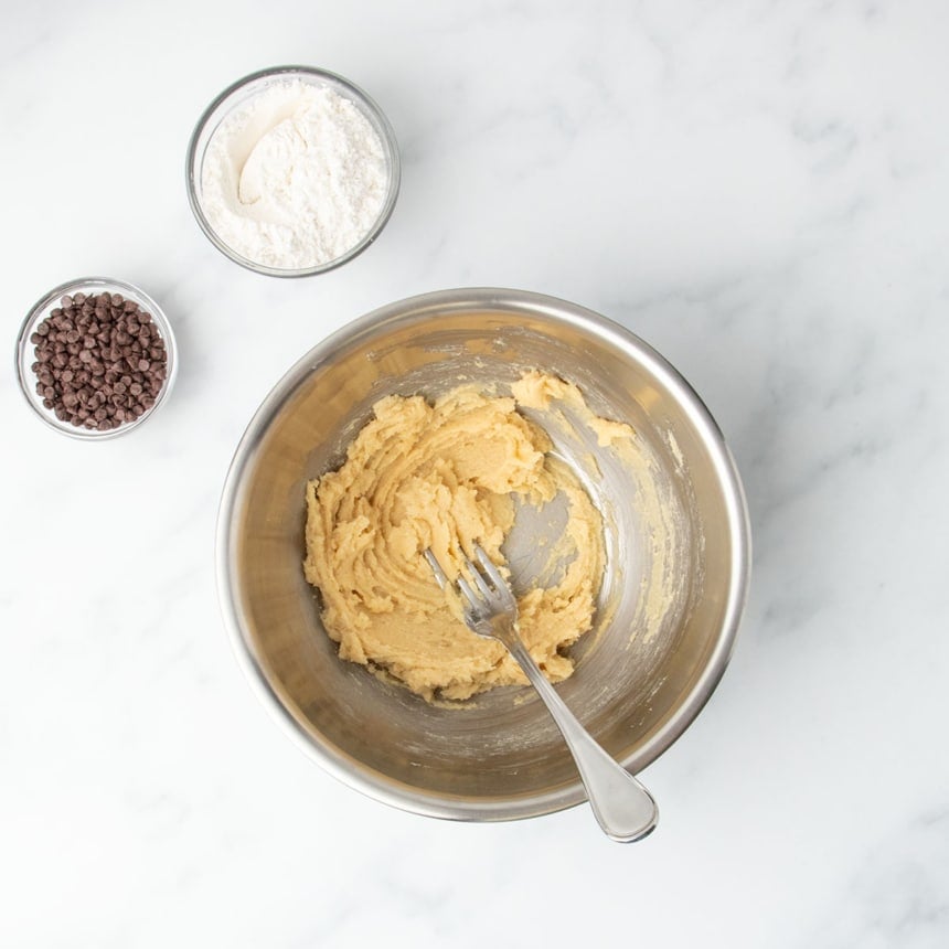 Smooth, pale light brown mixture of soft butter and sugars in a bowl, with gluten free flour and mini chocolate chips off to the side.