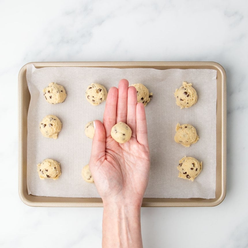 A single portion of shaped round of cookie dough held up in the palm of a hand, with other unrounded scoops on the tray below.